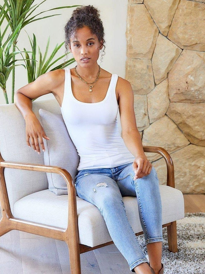 CAL JUNE white scoop neck tank top on a woman sitting in a chair, styled with ripped jeans and a necklace.
